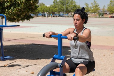 Azimli sporcu kadın açık hava spor aletleri üzerinde kürek çekiyor, sağlıklı yaşam tarzını ve formülünü destekliyor.
