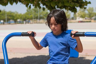 Açık hava spor salonunda mavi bir makinede egzersiz yapan konsantre bir çocuk.