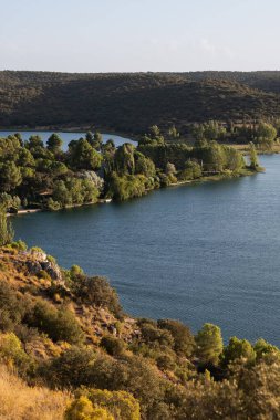 Mavi göl suyu ışığı yansıtıyor, yemyeşil ağaçlar ve açık gökyüzünün altında yuvarlanan tepelerle çevrili.
