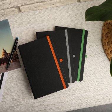 Turuncu, gri ve yeşil yer imli siyah defterler bir kalem ve bir derginin yanındaki beyaz bir masanın üzerinde duruyor.