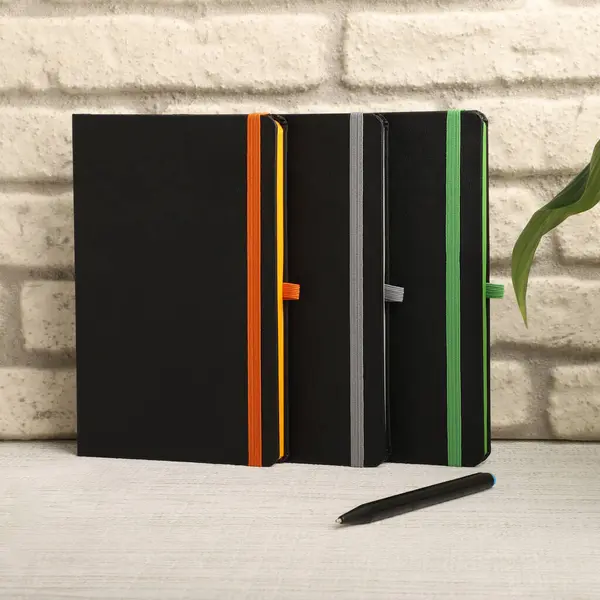 Siyah defterler, turuncu, gri ve yeşil elastik bantlarla tuğla bir duvara karşı ayakta duran siyah bir kalem eşliğinde.