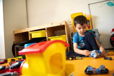 Genç çocuk renkli bir oyun odasında oyuncaklarla düşünceli bir şekilde meşgul oluyor.