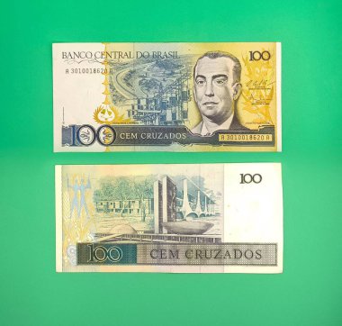 Brezilya 'dan gelen iki yüz bin cruzeiro banknotunun resmi, biri bir adamın portresi, diğeri ise modern bir binayı tasvir ediyor. Banknotlar canlı yeşil bir yüzeyde sergileniyor ve Brezilya 'nın ekonomik tarihini vurguluyor..