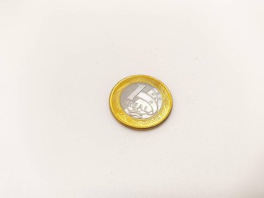 Tek hakiki bir para beyaz bir yüzeye dayanır. Metalik renkleri ve karmaşık detayları vurgular. Görüntü Brezilya parasının dokusunu ve şeklini yansıtıyor, ekonomi ve finansmanı simgeliyor..