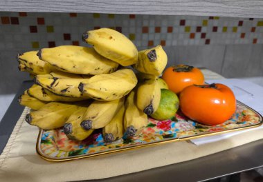 Bir demet sarı muz, iki portakal mandalinası ve yeşil limon içeren, canlı ve iştah açıcı bir kompozisyon oluşturan dekoratif bir tabakta güzel bir meyve düzenlemesi..