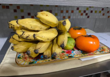 Tepside güzel bir taze meyve aranjmanı. Sarı muz, yeşil limon ve iki portakallı hurma var. Canlı renkler, nötr mutfak arka planıyla tezat oluşturarak tazelik ve sağlık katıyor..