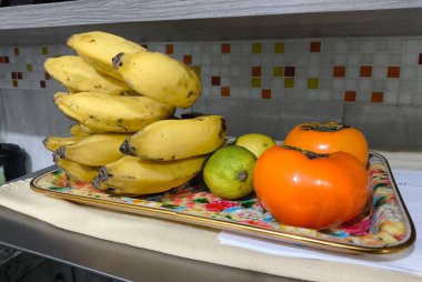 Canlı portakal hurmaları ve yeşil limonların yanında dekoratif tepside bir yığın sarı muz bulunan taze meyvelerden oluşan güzel bir düzenleme. Renkli fayanslarla aydınlık bir mutfak yüzeyindeki yerleşim neşeli ve davetkar bir imaj yaratıyor.