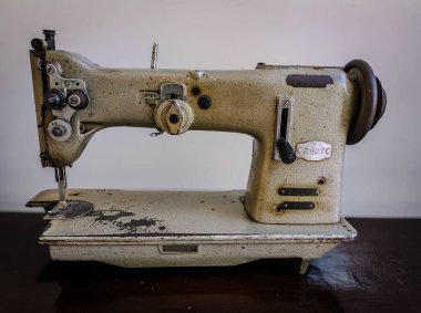 Eski bir dikiş makinesi, yıpranmış detaylarla nötr bir tonda tamamlanmış, parlak ahşap bir yüzeye yerleştirilmiş. Odak noktası yaşın güzelliği, nostaljiyi çağrıştırması ve dikiş sanatıyla olan bağlantıdır..