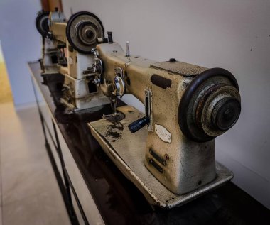 Minimalist bir ortamda sergilenen eski dikiş makinelerinin yakın çekimi. Görüntü, yıpranmış metal yapı ve ayarlama düğmeleri gibi ayrıntıları yakalıyor, nostalji ve işçiliği çağrıştırıyor..