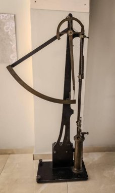 Modern bir ortamda sergilenen zarif ve klasik bir ölçüm aleti, muhtemelen bir teodolit veya seviye. Parlak metal ışığı yansıtırken kıvrımlar ve düz çizgiler ilginç bir kontrastlık yaratır. Tarafsız arkaplan nesneyi güçlendirir