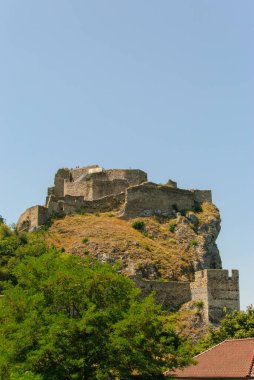 Antik şato kalıntıları, açık mavi gökyüzünün altında yemyeşil bir bitki örtüsüyle çevrili bir tepenin üzerinde oturur ve çarpıcı bir manzara yaratır.