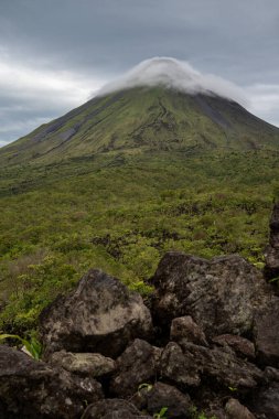 Pofuduk bulutlar ve yemyeşil manzaralarla çevrili görkemli bir volkanın çarpıcı manzarası.