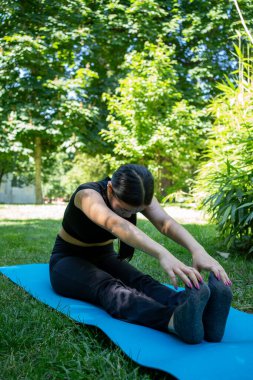 Bir kadın açık hava ortamında yoga yapıyor. Yeşilliklerle çevrili bir paspasın üzerinde zarifçe geriniyor.