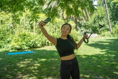 Bir kadın açık havada egzersiz yaparken canlı bir selfie çeker. Çevresi yemyeşil ve doğa ile doludur.