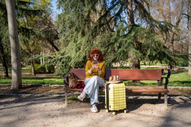 Şık ve kaygısız bir gezgin bir parkta rahat rahat oturuyor, yanında bir bavulla doğanın tadını çıkarıyor.