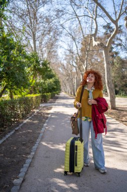 Güneşli bir parkta elinde sarı bir bavulla duran bir kadın seyahat ve maceranın özünü yakalıyor..