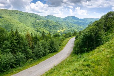 Yazın dağ manzarası boyunca yol. Yukarıdan manzara. Bulutlu gökyüzünün altında, benekli ışıkta yeşil doğa manzarası. Ormanların arasında kıvrılan güzel vadi
