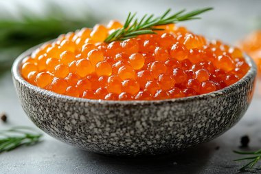 Yakından bakınca canlı turuncu somon yumurtası dolu bir kase görünüyor. Taze biberiyeyle süslenmiş, dokulu gri bir zemin üzerinde doğal zarafeti vurguluyor..