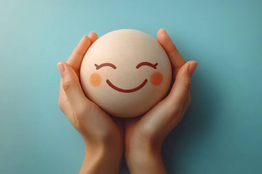 Rahatlatıcı bir stüdyo çekiminde iki el nazikçe neşeli bir çizgi film yüzü olan küreyi tutuyor, pembe yanaklarla büyütülmüş, dingin mavi bir arkaplan üzerine kurulmuş.