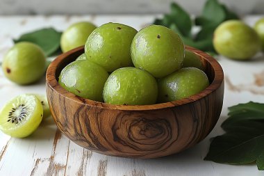 Görüntü, taze, parlak yeşil amla bektaşi üzümlerinin dokuma ahşap bir kase içinde taştığını gösterir; sahne yakınlarda canlı yeşil yapraklar ile sıkıntılı beyaz bir yüzey üzerine kuruludur.