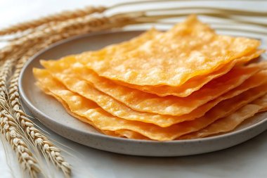 Yuvarlak bir tabağa konmuş yüksek açılı kızartılmış wonton ambalajları, beyaz bir yüzeye karşı dizilmiş, buğday sapları yakınlarda sıralanmış, doğal bir element eklenmiş..