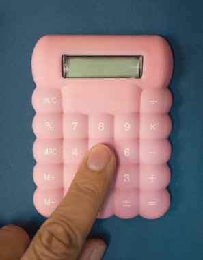 Mavi arkaplandaki pembe hesap makinesindeki bir sayı düğmesine basan parmaklar, muhasebe, finans ve matematik konularını canlı ve çekici bir şekilde resmediyor..