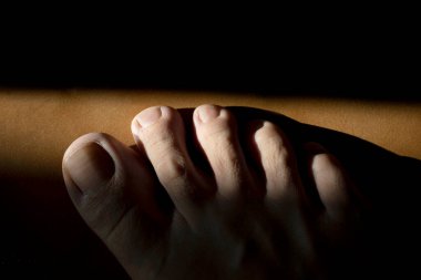 Kısmen aydınlanmış ayak parmakları kahverengi bir yüzeyde duruyor, karanlıktan bir ışık şeridine dönüşüyor, çarpıcı bir kontrastlık yaratıyor ve derinin dokusunu parlaklaştırıyor.