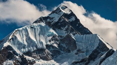Karla kaplı bir dağ zirvesi mavi bir gökyüzüne karşı dimdik duruyor. Dağılmış bulutlar görkemli bir manzara sergiliyor..
