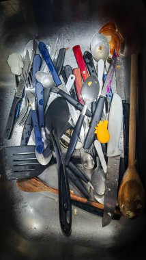 Çatallar, bıçaklar, kaşıklar ve spatulalar dahil mutfak aletleriyle dolu bir lavabo. Çatal bıçaklar birbirine dolanmış, bazıları kirli, yağ izleri ve yiyecek kalıntılarıyla, mutfakta kargaşa ve sıkı çalışma ortamı yaratıyor..