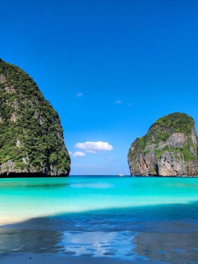 Plajda güneşli bir gün. Arkasında kaya oluşumları ve bitki örtüsü olan berrak turkuaz bir su. Maya Körfezi, Tayland.