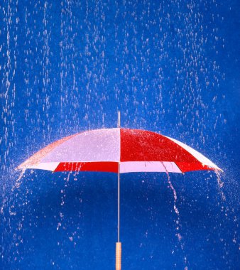 Düşen suya karşı canlı kırmızı ve pembe çizgili şemsiye koruyucu, canlı mavi arka plana karşı görsel bir kontrastlık yaratır, yağmurlu havalarda korunma ve direnci simgeler..