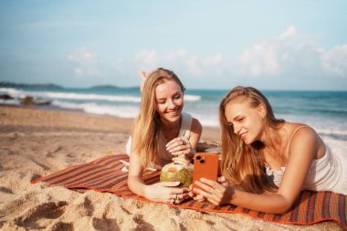Genç kadınlar plaj atmosferinin tadını çıkarıyorlar, içkilerini yudumluyorlar ve güneşin altında telefonlarını kaydırıyorlar. Yüksek kalite fotoğraf