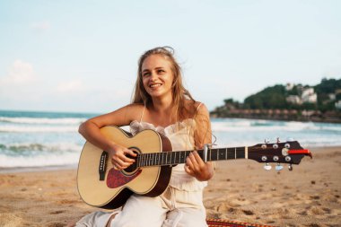 Sahilde gitar çalan, gün batımında ıslanan ve okyanusun rahatlatıcı seslerini dinlendiren bir kadın. Yüksek kalite fotoğraf