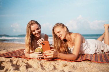 Genç kadınlar güneşli bir plaj gününün tadını çıkarırlar, içkilerini yudumlarlar ve anılarını yakalamak için akıllı telefonlarını kullanırlar. Yüksek kalite fotoğraf