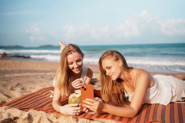 İki arkadaş plajda dinlenir, akıllı telefonlarıyla anılarını yakalar ve sıcak güneşin altında içkilerinin tadını çıkarırlar.