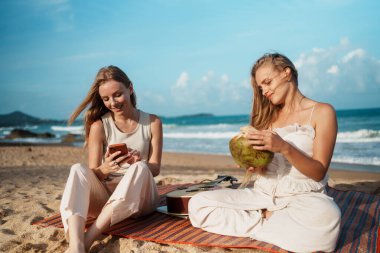 Kumsalda eğlenceli ve rahatlatıcı bir gün. Genç kadınlar gülümsüyor, içkilerinin tadını çıkarıyor ve akıllı telefonlarıyla bağlantıda kalıyorlar. Yüksek kalite fotoğraf