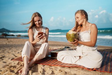 Kumsalda eğlenceli ve rahatlatıcı bir gün. Genç kadınlar taze hindistan cevizinin tadını çıkarırken ve akıllı telefonlarıyla bağlantıda kalırken. Yüksek kalite fotoğraf