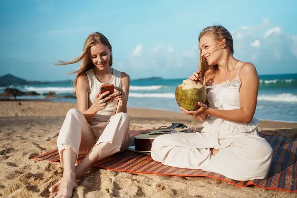 Kumsalda eğlenceli ve rahatlatıcı bir gün. Genç kadınlar taze hindistan cevizinin tadını çıkarırken ve akıllı telefonlarıyla bağlantıda kalırken. Yüksek kalite fotoğraf