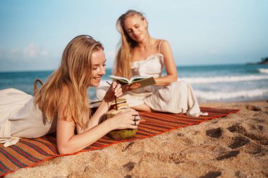 Güneşli bir sahil gününde, iki arkadaş, deniz kenarında dinlenirken ve kitap okurken hindistan cevizi içkisi yudumlarlar.
