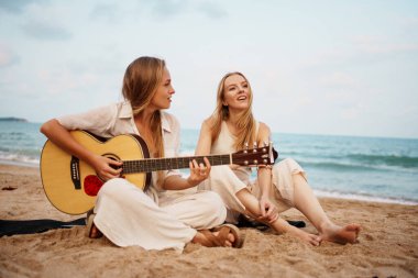 Bir grup arkadaş, huzurlu plaj ortamında gitar çalarken neşeli ve kaygısız anları paylaşıyorlar.