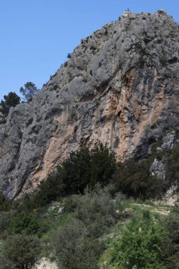 Fotoğraf, parçalı bulutlu bir gökyüzünün altında engebeli, kayalık tepeleri olan dağlık bir manzarayı gösteriyor. Dağlar ise dipdiri bir bitki (ler) ve hoş kokulu ağaçlarla kaplıdır. Ön planda..
