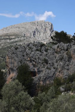 Fotoğraf, parçalı bulutlu bir gökyüzünün altında engebeli, kayalık tepeleri olan dağlık bir manzarayı gösteriyor. Dağlar ise dipdiri bir bitki (ler) ve hoş kokulu ağaçlarla kaplıdır. Ön planda..