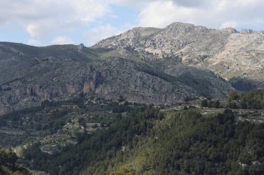 Fotoğraf, parçalı bulutlu bir gökyüzünün altında engebeli, kayalık tepeleri olan dağlık bir manzarayı gösteriyor. Dağlar ise dipdiri bir bitki (ler) ve hoş kokulu ağaçlarla kaplıdır. Ön planda..