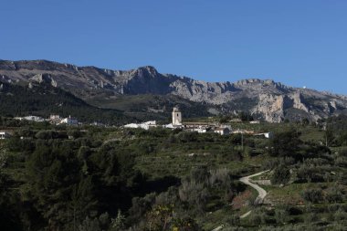 Fotoğraf, parçalı bulutlu bir gökyüzünün altında engebeli, kayalık tepeleri olan dağlık bir manzarayı gösteriyor. Dağlar ise dipdiri bir bitki (ler) ve hoş kokulu ağaçlarla kaplıdır. Ön planda..