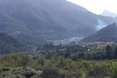 Fotoğraf, parçalı bulutlu bir gökyüzünün altında engebeli, kayalık tepeleri olan dağlık bir manzarayı gösteriyor. Dağlar ise dipdiri bir bitki (ler) ve hoş kokulu ağaçlarla kaplıdır. Ön planda..