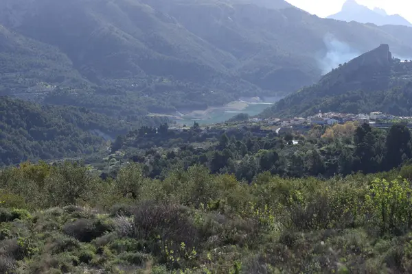 Fotoğraf, parçalı bulutlu bir gökyüzünün altında engebeli, kayalık tepeleri olan dağlık bir manzarayı gösteriyor. Dağlar ise dipdiri bir bitki (ler) ve hoş kokulu ağaçlarla kaplıdır. Ön planda..