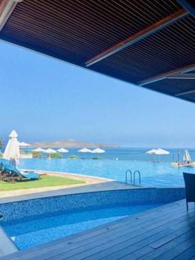 Lüks bir tatil köyünde denize bakan sonsuz bir havuzu gösteren sakin bir sahne. Şemsiyeler ve şezlonglar mavi gökyüzünün altında, rahatlama ve tatil hissi uyandırıyor..