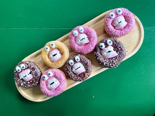 Komik ve ürkütücü cadılar bayramı temalı çörekler şeker gözlü ve sahte dişlerle süslenmiş, yeşil bir masanın üzerindeki tahta tepsiye yerleştirilmiş. Cadılar Bayramı partisi atıştırmalıkları ya da yaratıcı pişirme fikirleri için mükemmel..