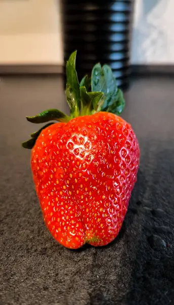 Siyah bir masanın üstünde bir çilek. Çileklere yakın çekim. Arka planda beyaz bir duvar ve siyah metal bir teneke kutu..