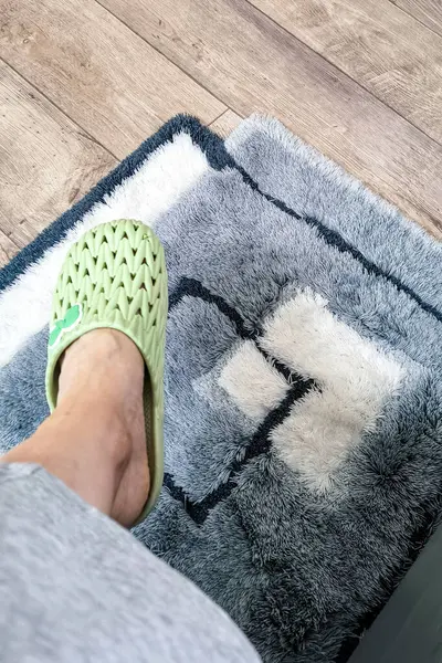 Ahşap zeminde geometrik desenli yumuşak gri ve beyaz kabarık bir kilimin üzerinde yeşil bir terlik giymiş bir kadın ayağı. Samimi minimalist oda iç görüntü ve yaşam tarzı konsepti.